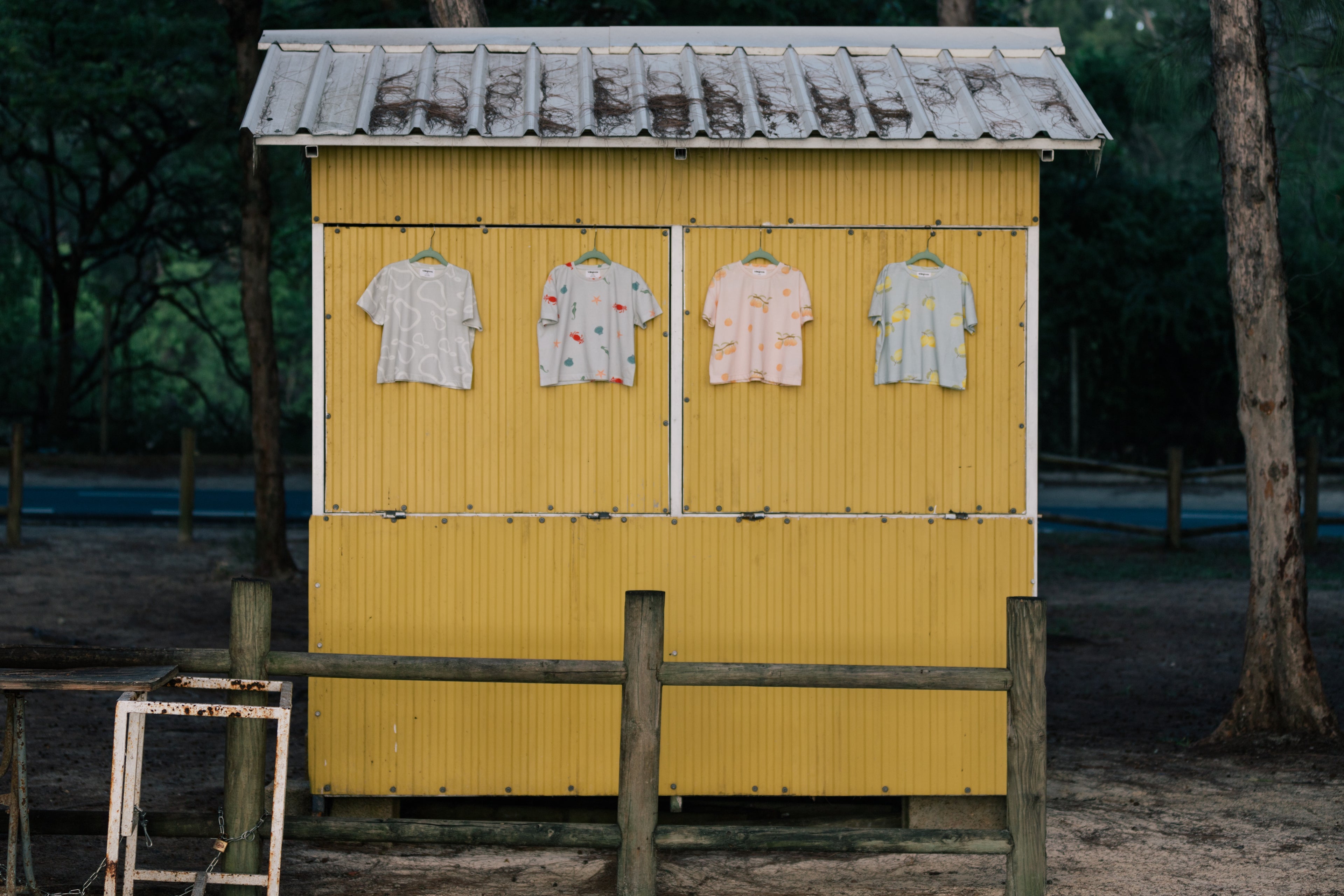 Wildgreen’s Island Life collection featuring four organic cotton T-shirts—White Pear, Marine, Apricot, and Lemon—hanging on a tropical island fruit shack. Ethically made and gender-neutral, these sustainable kids’ tees blend stylish prints with eco-conscious fashion. #OrganicKidswear #SustainableFashion