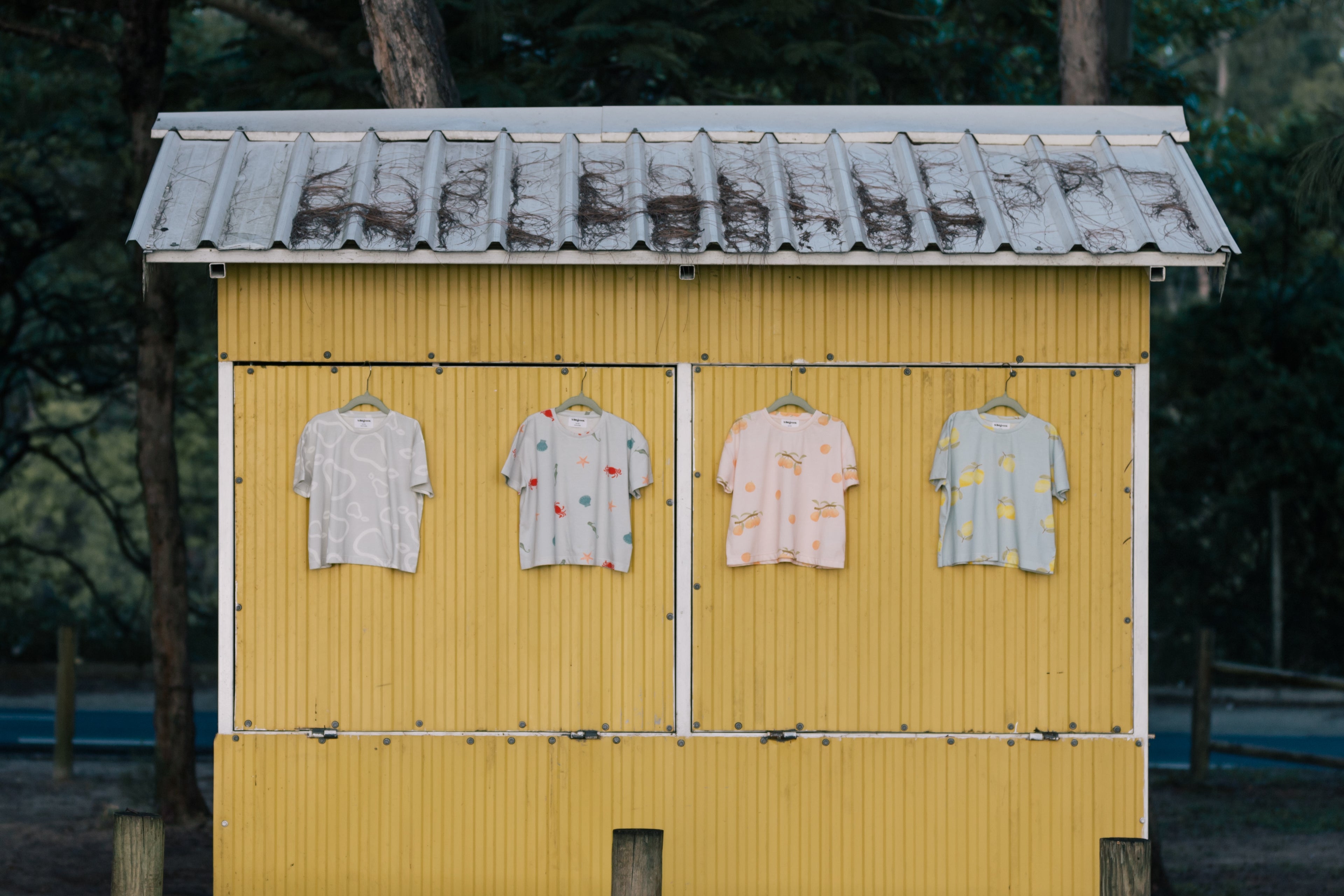 Wildgreen’s Island Life collection featuring four organic cotton T-shirts—White Pear, Marine, Apricot, and Lemon—hanging on a tropical island fruit shack. Ethically made and gender-neutral, these sustainable kids’ tees blend stylish prints with eco-conscious fashion. #OrganicKidswear #SustainableFashion